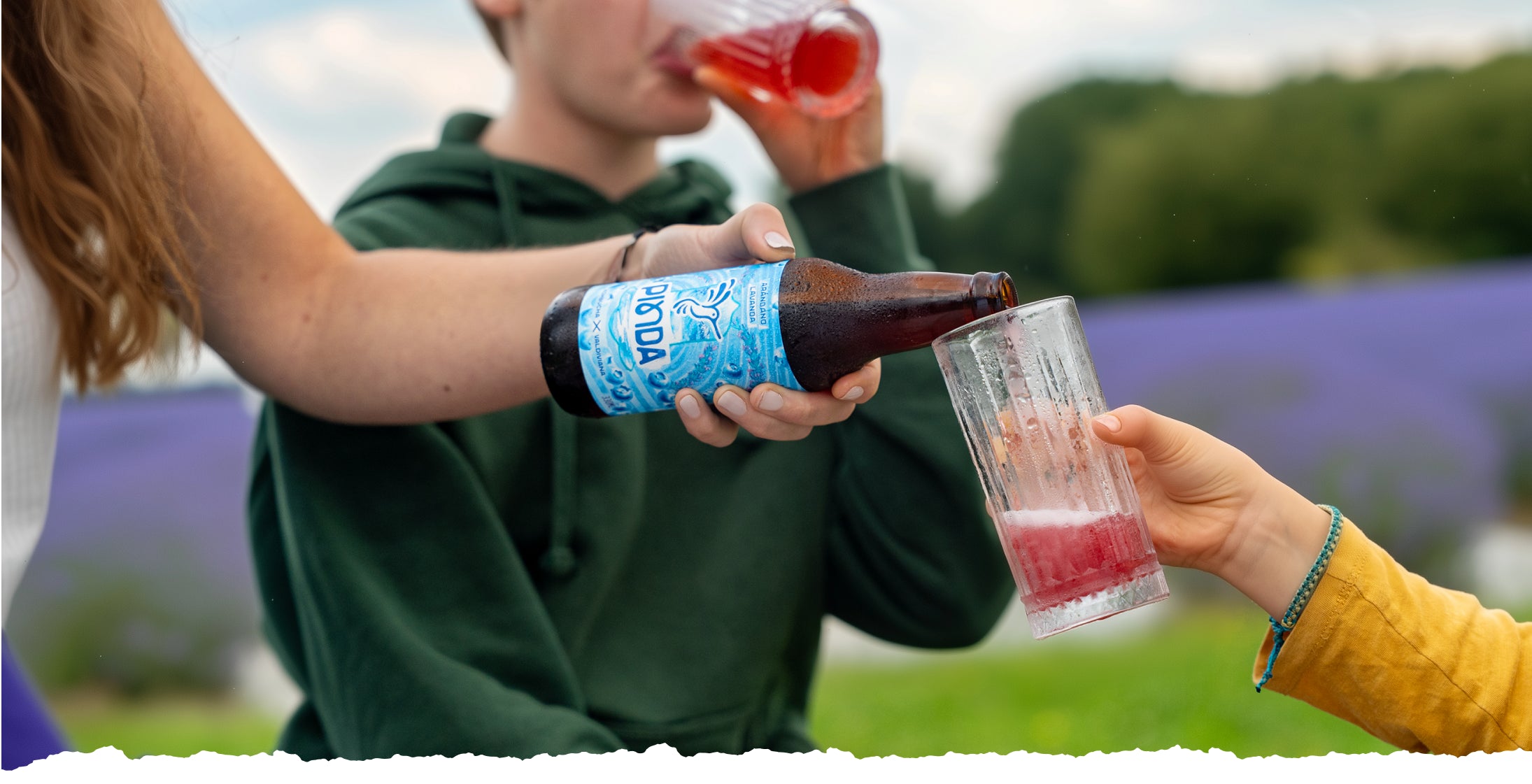Madre sirviendo kombucha Pinda a sus hijos en campo de lavanda al atardecer. Botellas artesanales con etiquetas azules, bebida burbujeante roja. Escena familiar de conexión y bienestar natural con super bebida probiótica.