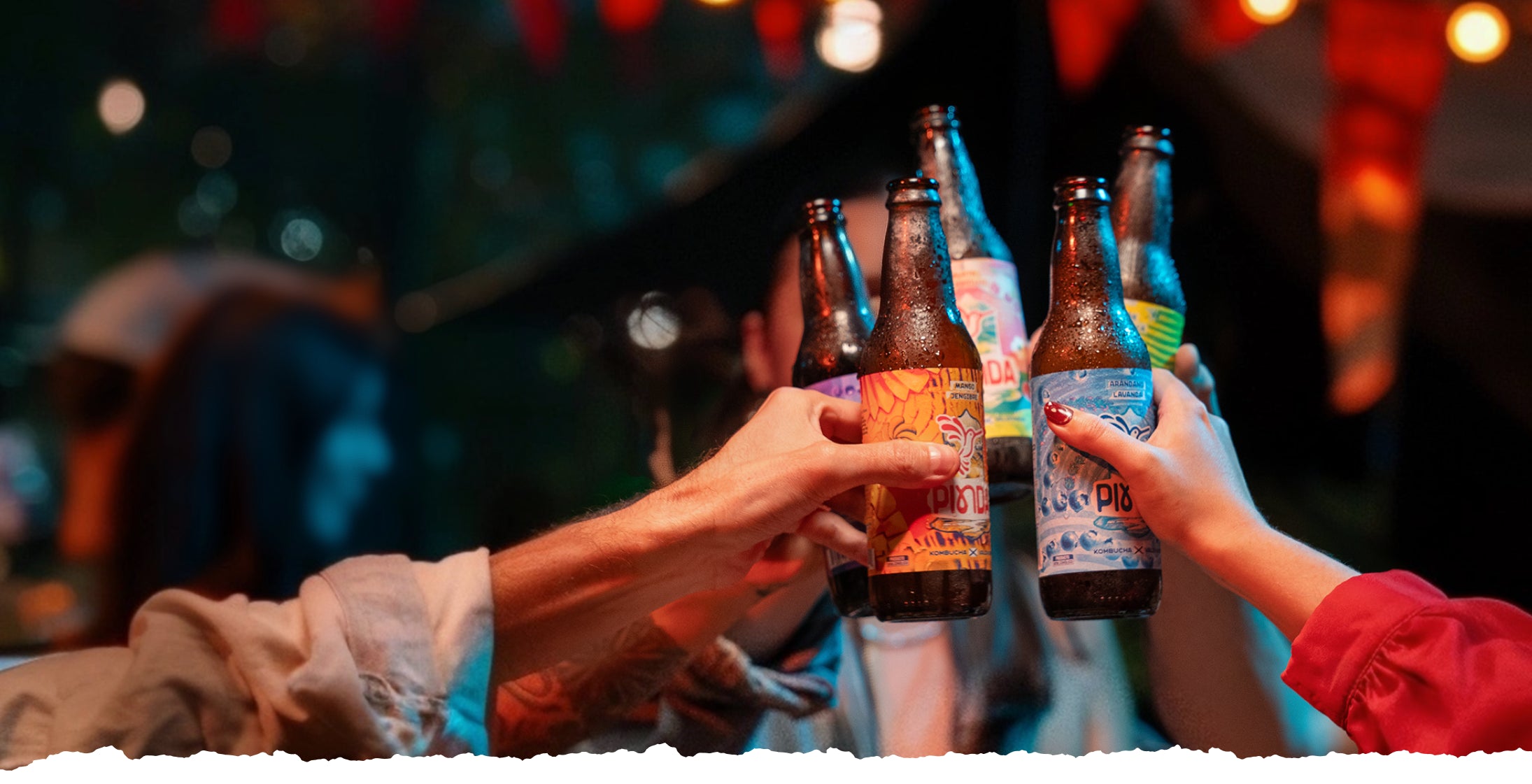 Grupo diverso brindando con kombucha PINDA en celebración nocturna al aire libre. Luces decorativas y botellas artesanales con etiquetas coloridas destacan la esencia de comunidad consciente y ritual de bienestar. Experiencia sensorial con super bebida probiótica de la Selva Valdiviana.