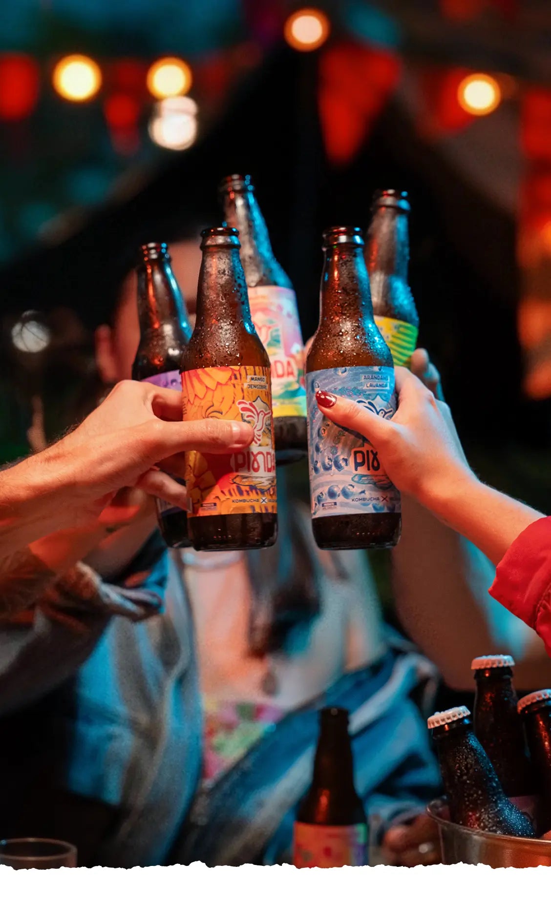 Celebración con kombucha PINDA: amigos brindan con bebida artesanal probiótica en noche con luces. Comunidad consciente y ritual de bienestar natural.